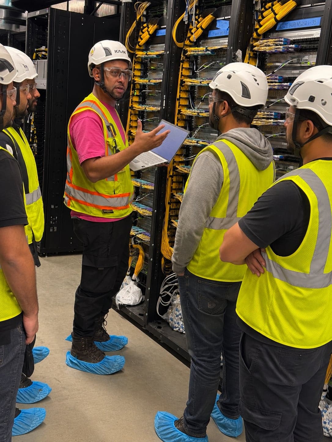 Kharroubi Fiber Service technician in a hard hat and safety vest holding a laptop and briefing four crew members beside fiber distribution racks in a data center